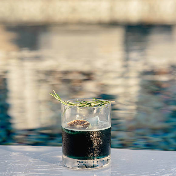 Grand Ambassador pool bar cocktail with ice, rosemary garnish, and poolside reflections