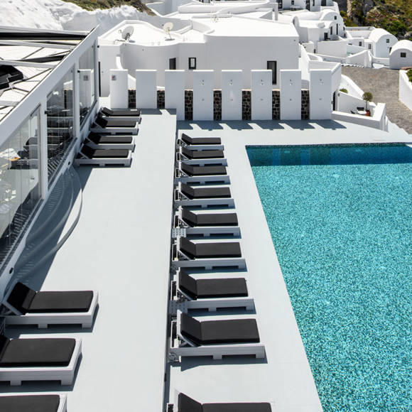 Grand Ambassador swimming pool with modern black sunbeds, glass walls, and whitewashed Santorini architecture