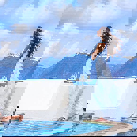 Man relaxing in pool and woman in flowing white dress standing by poolside with caldera cliffs and blue sky in background at Grand Ambassador