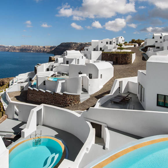 Aerial view of Grand Ambassador hotel showing white-washed buildings with private pools overlooking the deep blue sea under a partly cloudy sky