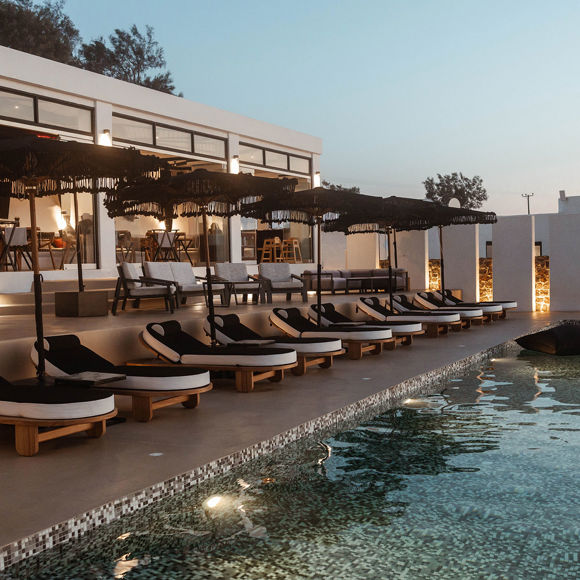 Grand Ambassador pool bar at dusk with illuminated pool, round sunbeds, parasols, and restaurant seating inside