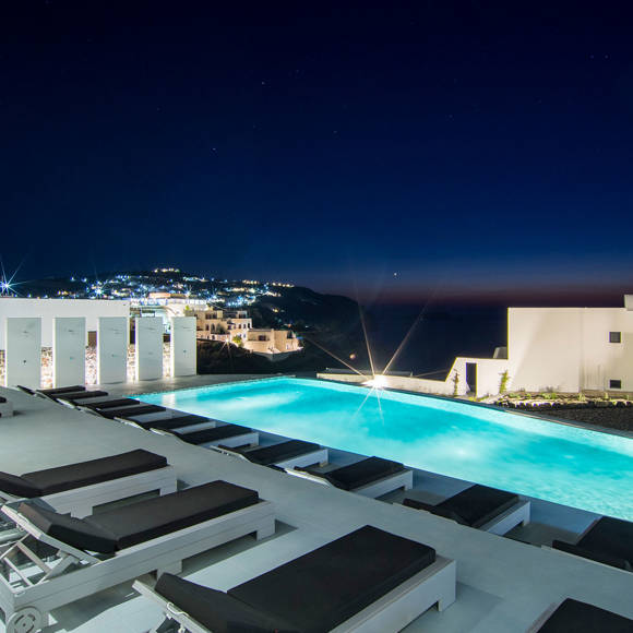 Grand Ambassador swimming pool illuminated at night with sunbeds and panoramic Santorini skyline view