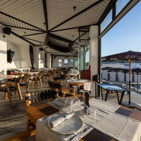 Grand Ambassador Dione restaurant interior with wooden chairs, set tables, and view of pool and umbrellas