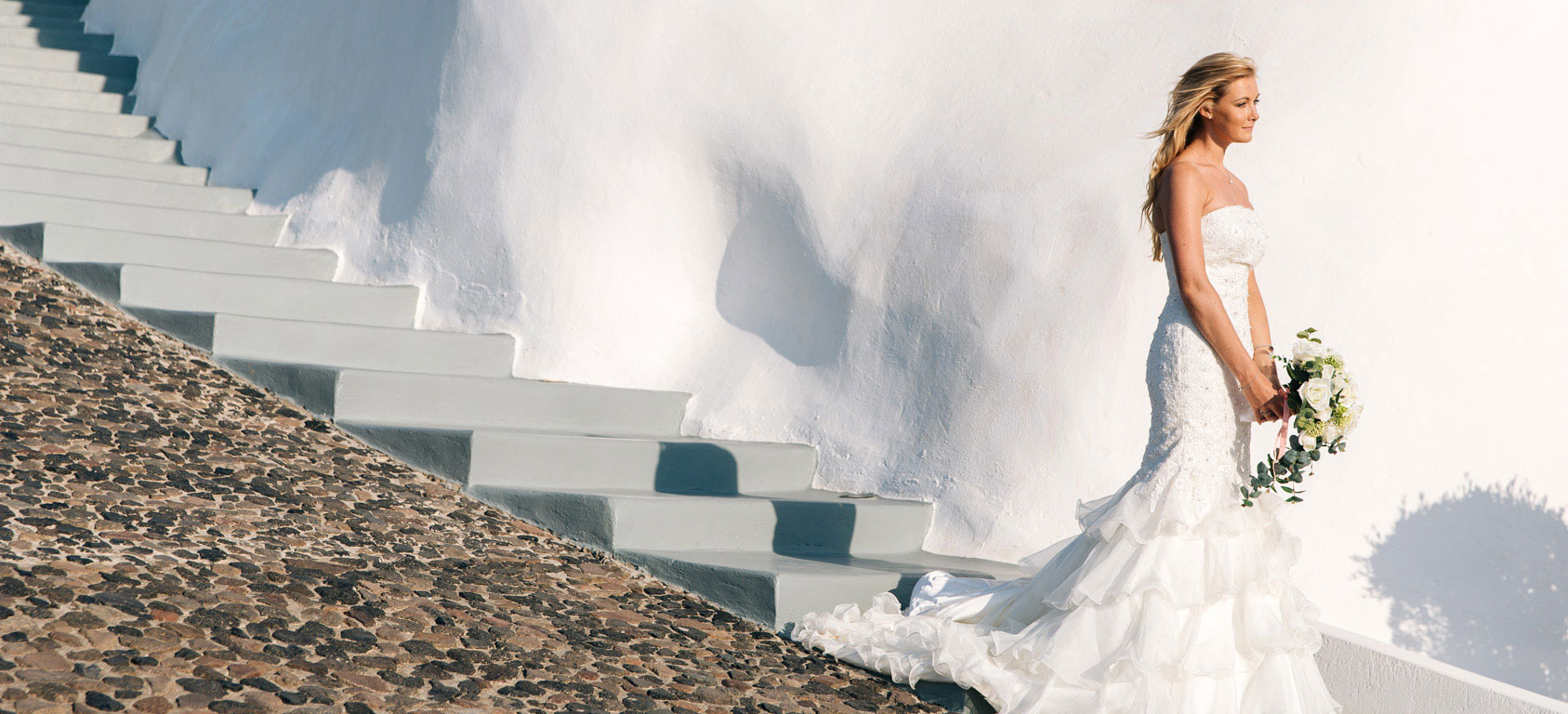 Bride in white wedding dress holding bouquet by white wall and stone stairs in sunlight at Grand Ambassador