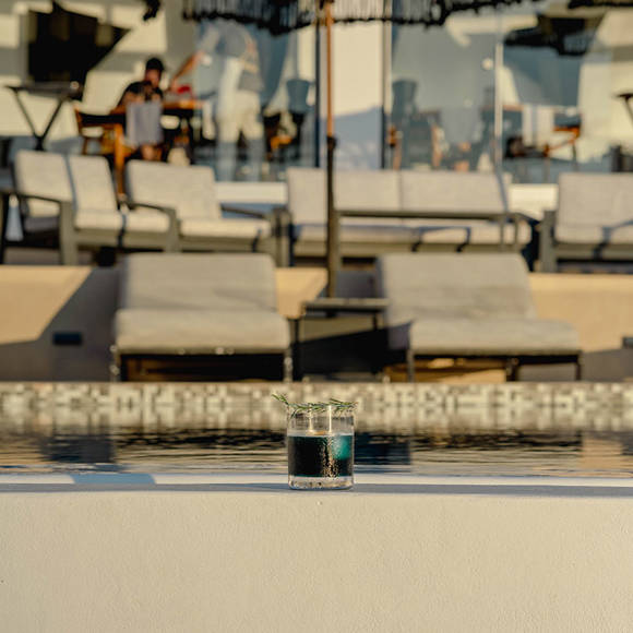 Grand Ambassador pool bar cocktail with rosemary garnish placed on pool edge, sunbeds and parasols in background