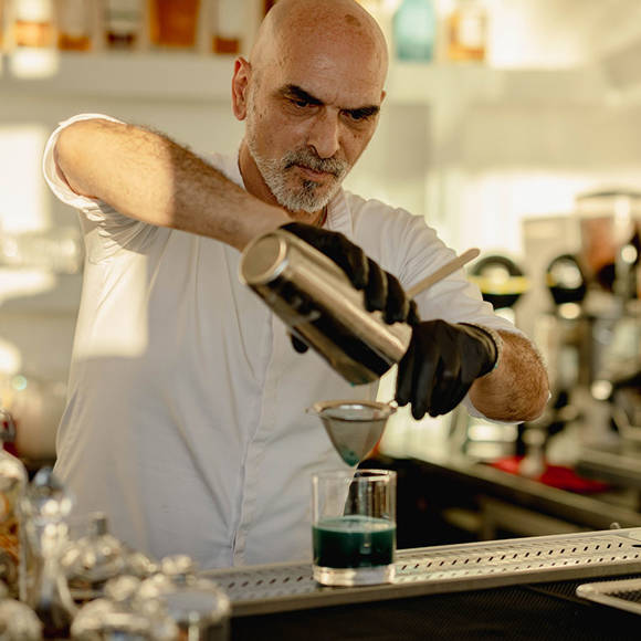 Grand Ambassador pool bar bartender wearing gloves pouring blue cocktail through strainer into glass with bottles in background