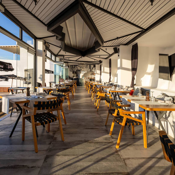Grand Ambassador Dione restaurant bright interior with modern wooden chairs, set tables, and glass walls overlooking pool area