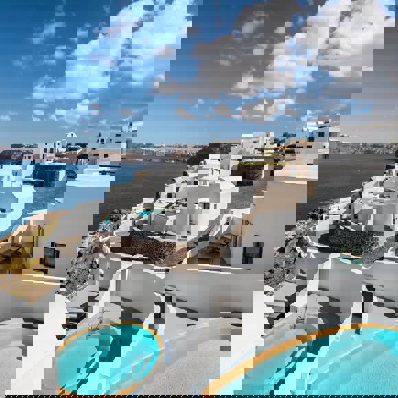 Outdoor view of Grand Ambassador hotel’s whitewashed villas perched on a cliff overlooking the blue sea, featuring private circular pools and a partly cloudy sky