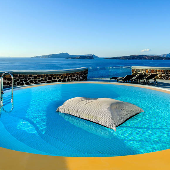 Round pool with floating cushion and sea view at Grand Ambassador in Santorini, overlooking volcanic islands under a clear blue sky
