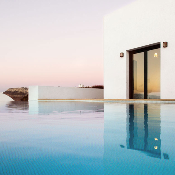Grand Ambassador Santorini: Infinity pool reflecting white modern building at sunset with volcanic cliffs backdrop