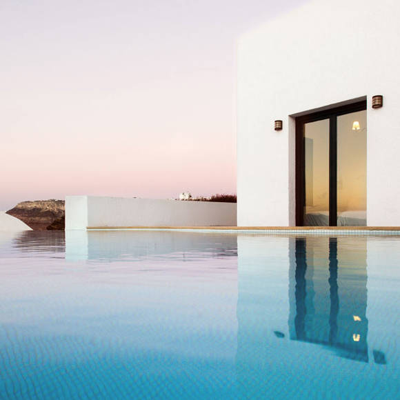 Grand Ambassador Santorini: Infinity pool reflecting white modern building at sunset with volcanic cliffs backdrop